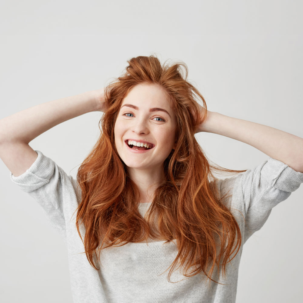 Lächelnde Person mit vollem, rotem Haar hält Hände am Kopf, symbolisiert Haargesundheit.